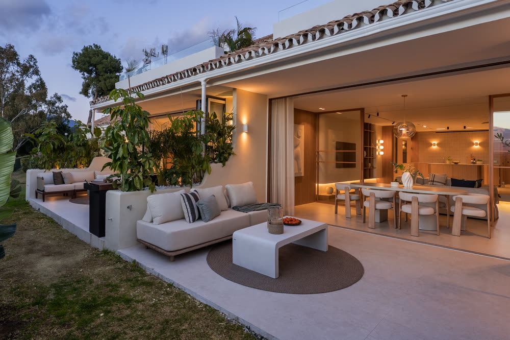 Covered terrace of a modern villa at dusk, lights on in the open-plan living room