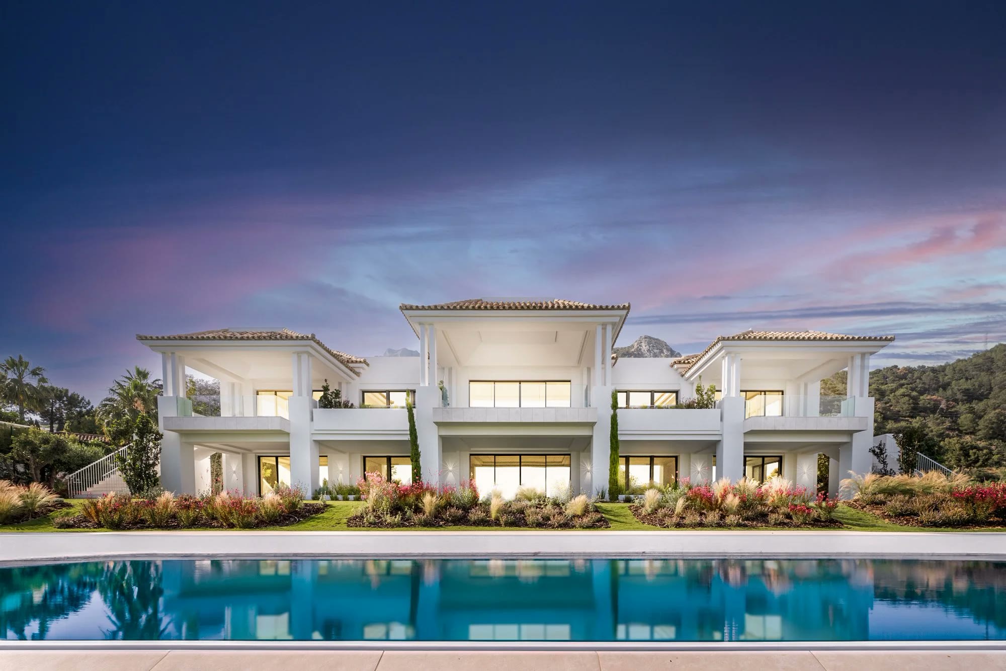 Row of white villas above a pool at dusk, lights on inside